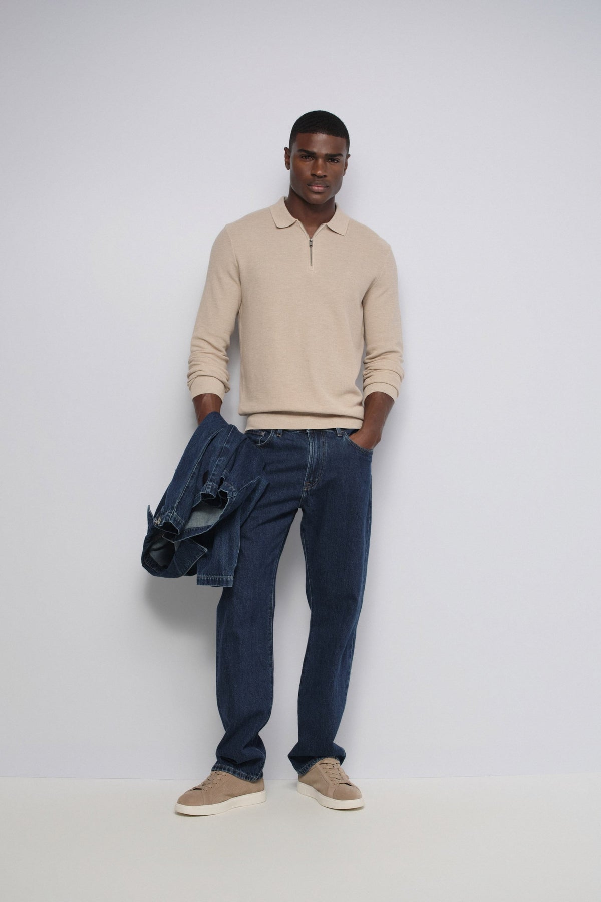 A man modeling a beige long-sleeve polo shirt, dark blue corduroy trousers, and beige slip-on shoes, holding a dark blue jacket against a plain light gray studio background.