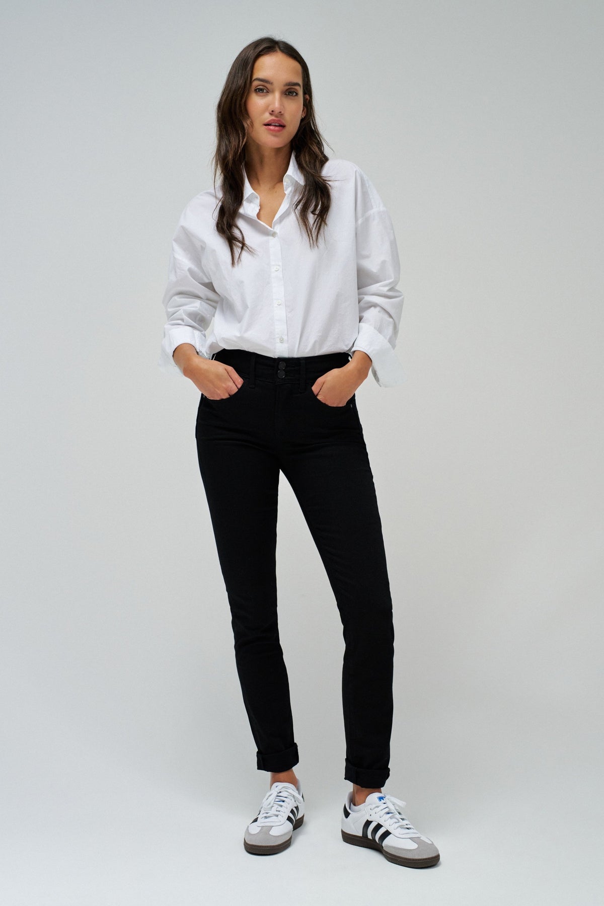 A woman modeling a white long-sleeve button-up shirt, black slim-fit jeans, and white sneakers with black stripes, standing against a light gray studio background.