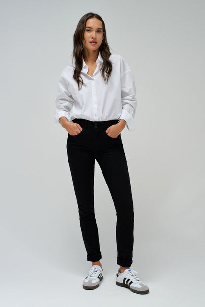 A woman modeling a white long-sleeve button-up shirt, black slim-fit jeans, and white sneakers with black stripes, standing against a light gray studio background.