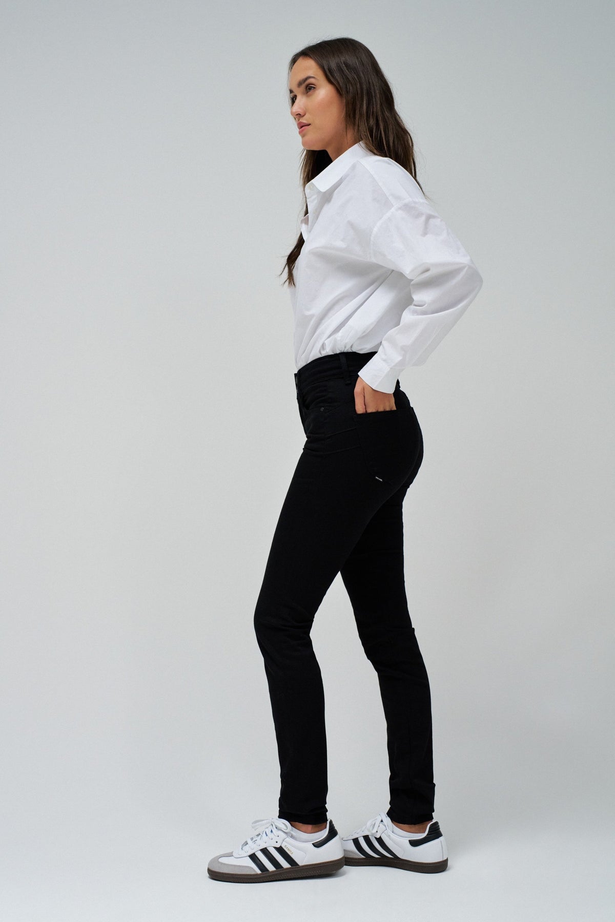 A woman modeling a white long-sleeve button-up shirt and black skinny jeans, paired with white sneakers featuring black stripes, set against a plain light gray studio background.
