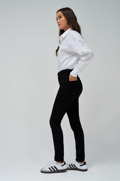 A woman modeling a white long-sleeve button-up shirt and black skinny jeans, paired with white sneakers featuring black stripes, set against a plain light gray studio background.