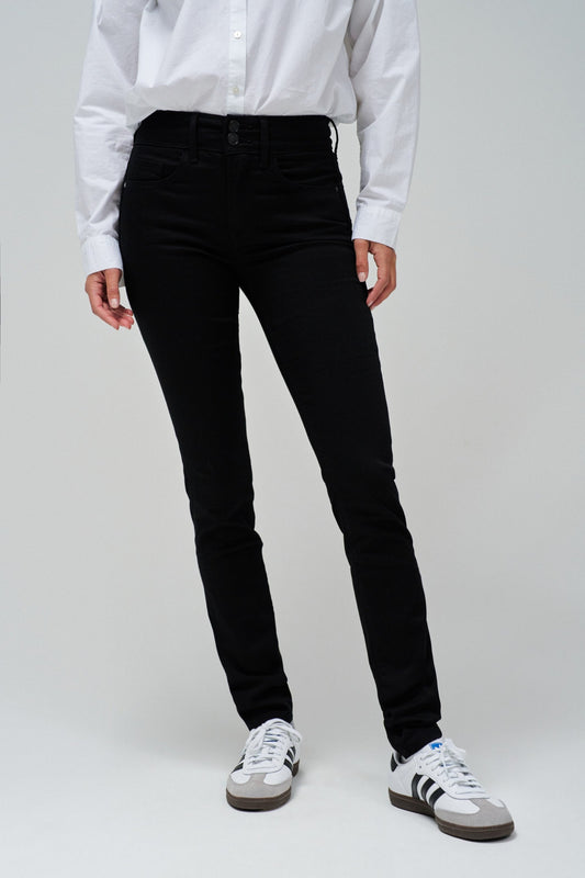 A model wearing black slim-fit jeans, a white long-sleeve button-up shirt, and white sneakers with black stripes, shown from the waist down against a light gray studio background.