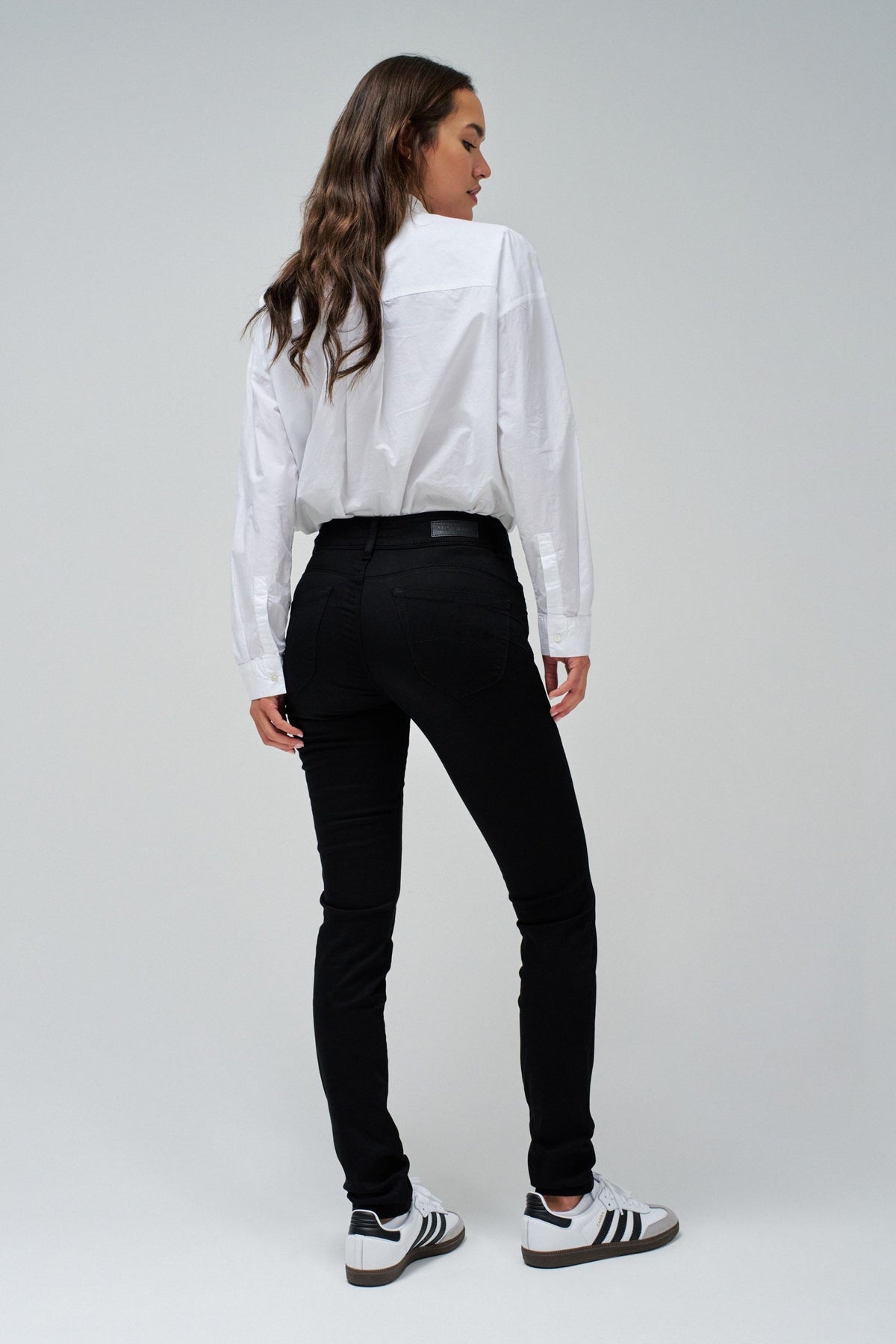 A woman is shown from the back wearing a white long-sleeve button-up shirt, black slim-fit jeans, and white sneakers with black stripes, set against a plain light gray studio background.