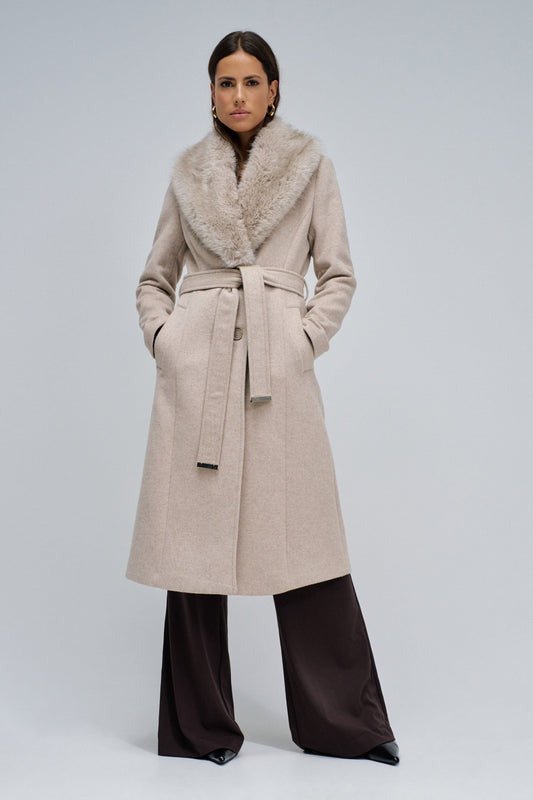 A woman models a long beige wool coat with a large beige fur collar, belted at the waist, paired with dark brown wide-leg trousers, against a light gray studio background.