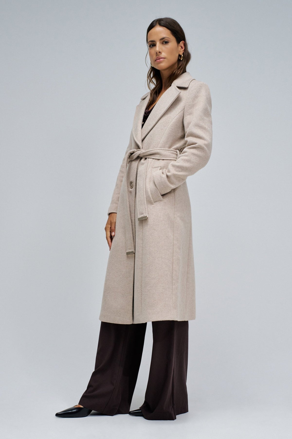 A woman modeling a long, light beige belted coat over dark wide-leg trousers, standing against a plain light gray studio background.