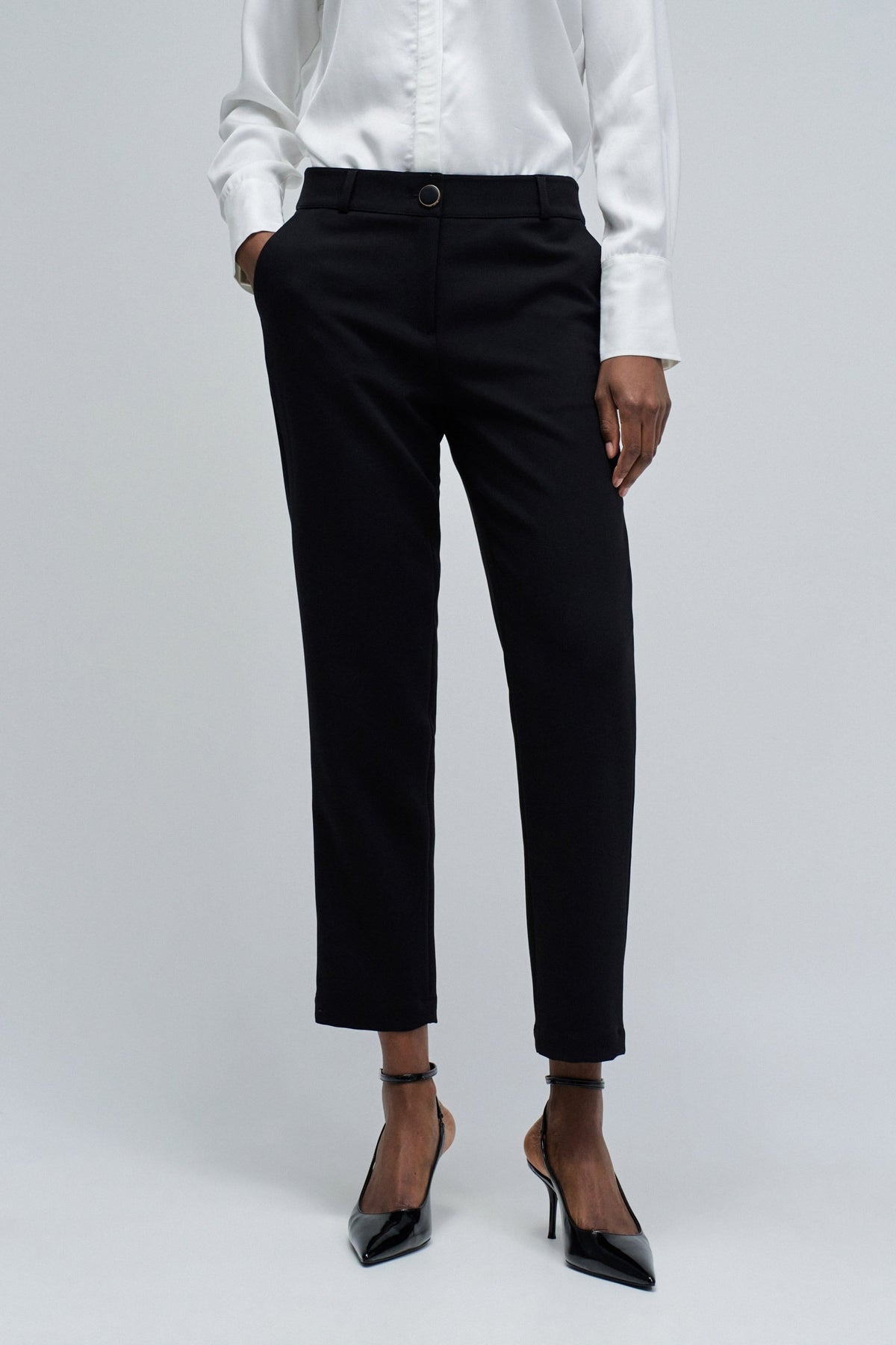 A model wearing black cropped trousers, a white long-sleeve blouse, and black pointed-toe heels, standing against a light gray studio background.
