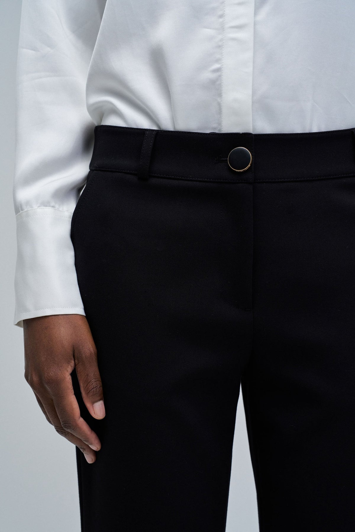 Close-up of a person wearing black tailored trousers and a white button-up shirt, showing the waistband with a single dark button.
