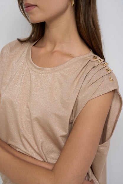 A close-up of a woman wearing a shimmering champagne beige top with gold button details on the shoulder, shown against a plain light background.