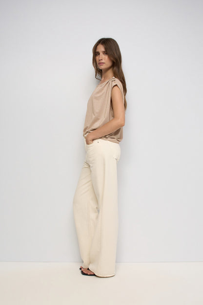 A woman stands in profile wearing a beige draped sleeveless top and wide-leg cream trousers against a plain light gray studio background.