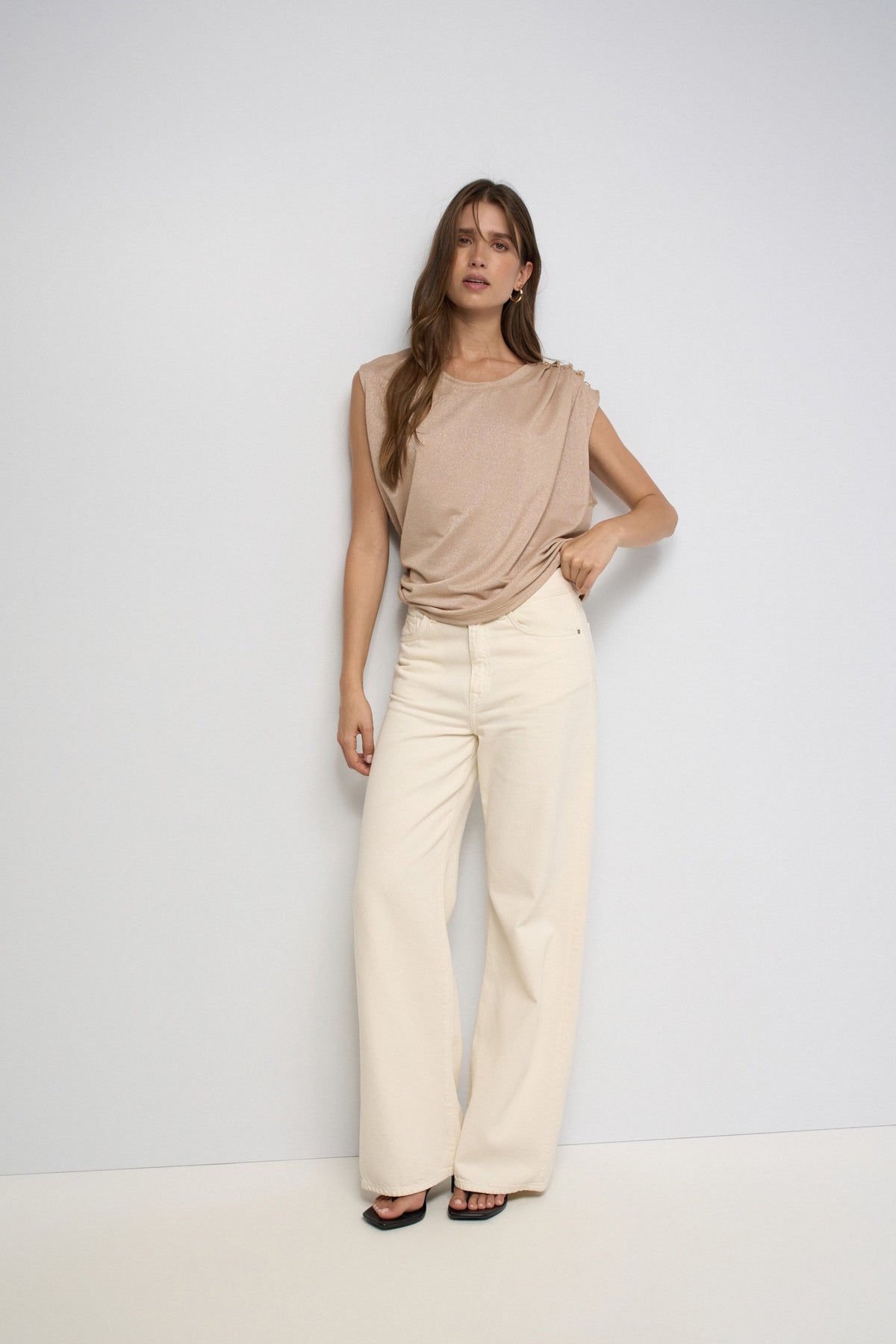 A woman modeling a sleeveless beige top and cream wide-leg trousers against a plain white studio background.