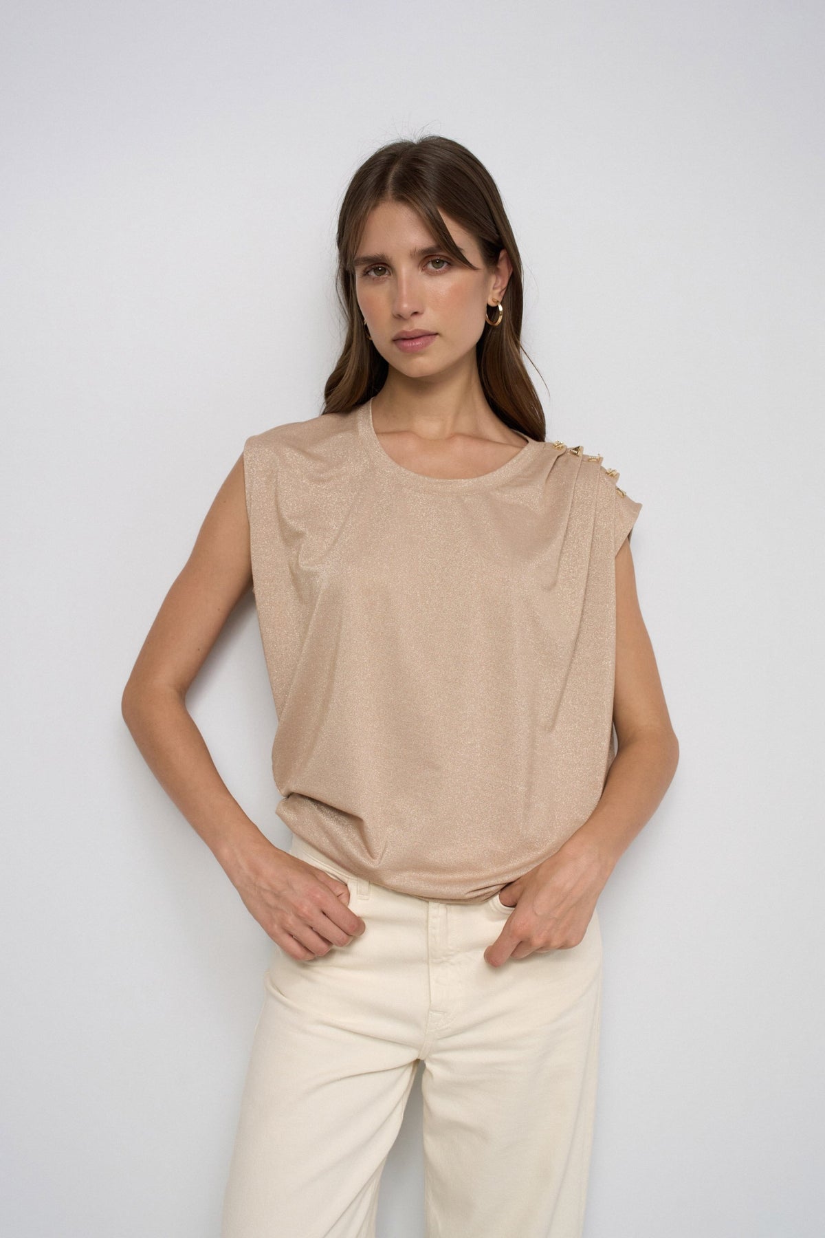 A woman modeling a sleeveless beige top with draped shoulder details and cream-colored pants, standing against a light gray studio background.