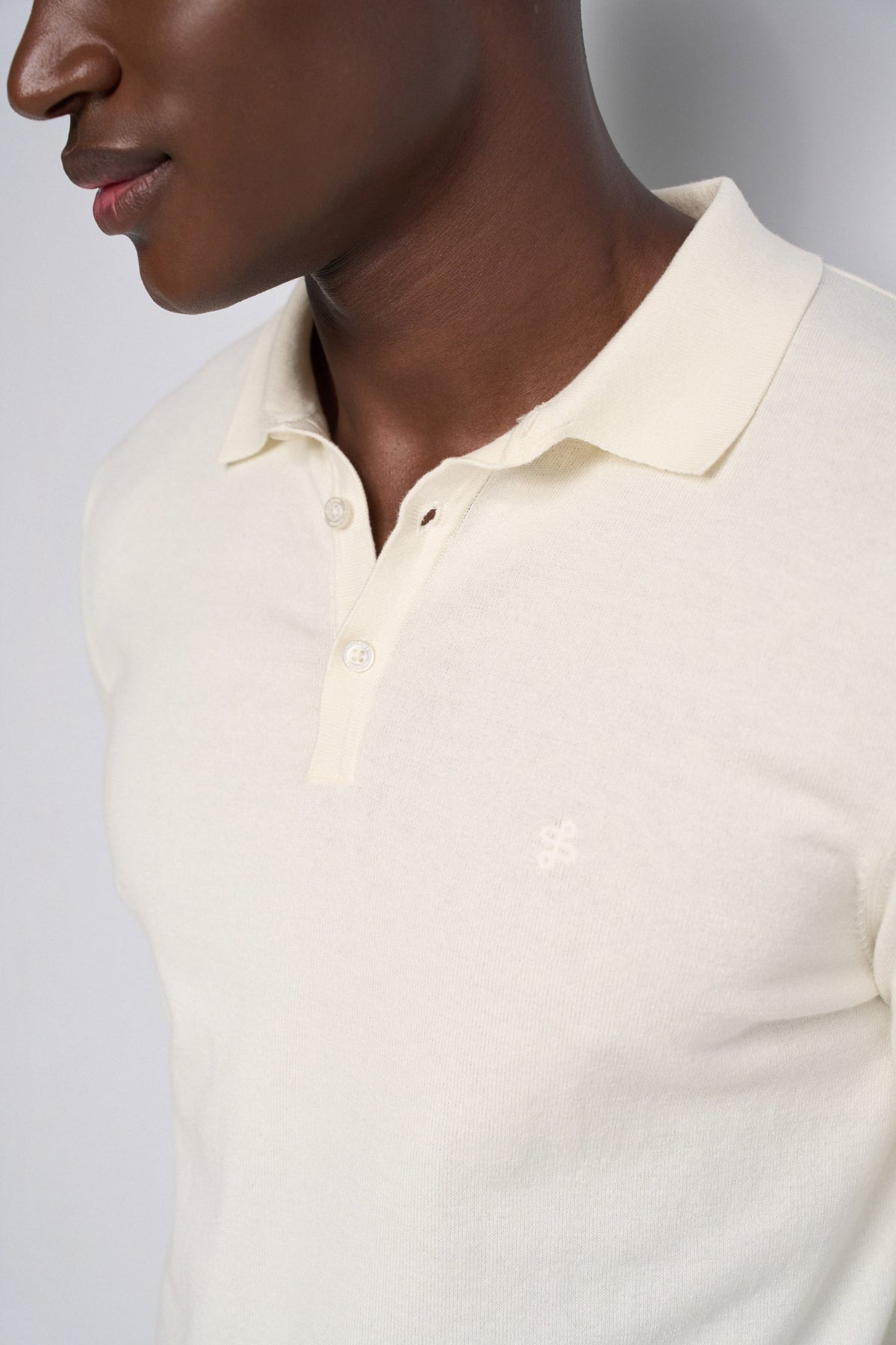 Close-up of a man wearing a cream-colored short-sleeve polo shirt with a small embroidered logo on the chest, set against a neutral gray background.