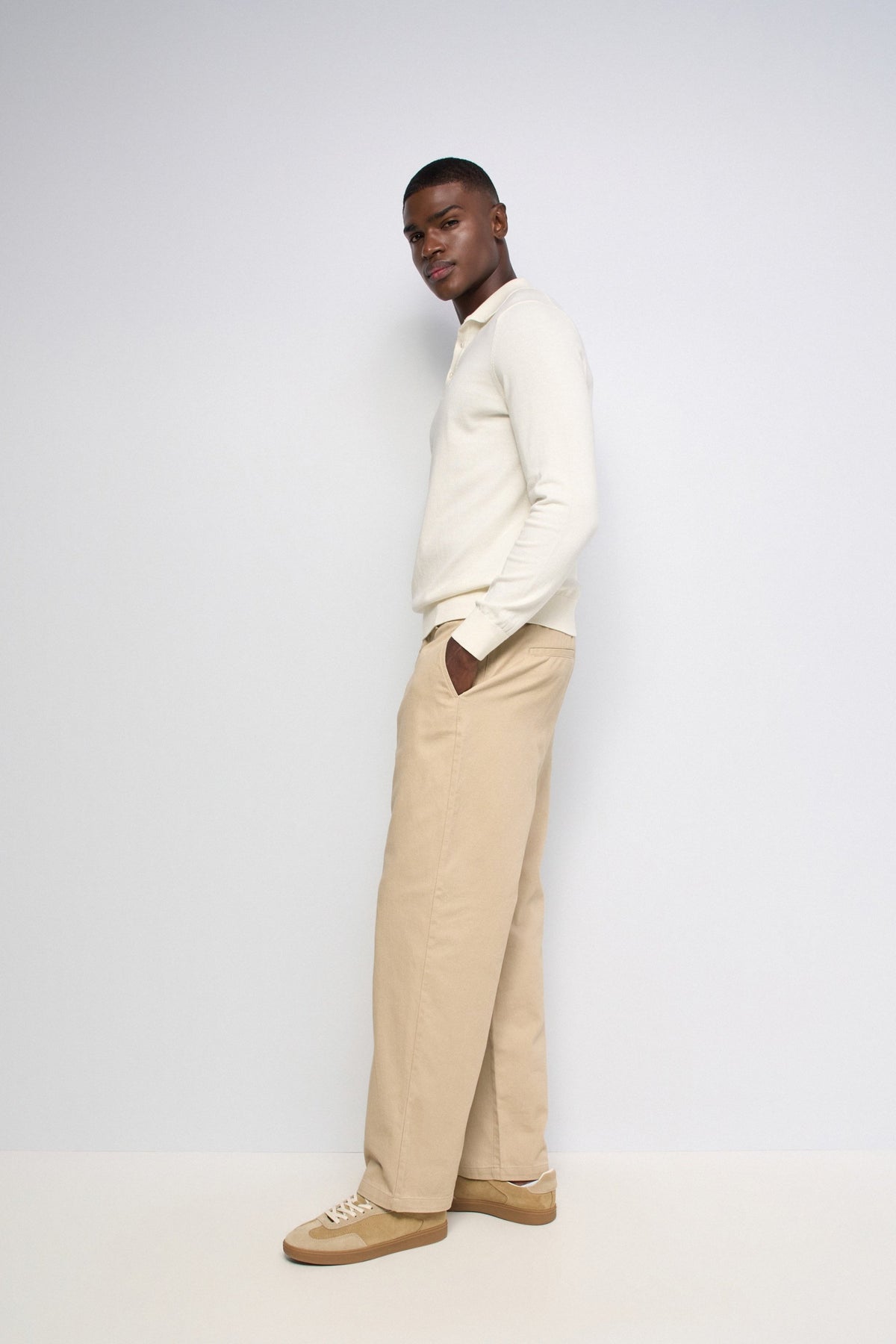 A man wearing a cream long-sleeve knit polo shirt, beige wide-leg trousers, and tan sneakers, standing in profile against a plain light gray studio background.