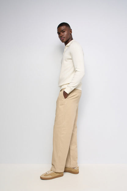 A man wearing a cream long-sleeve knit polo shirt, beige wide-leg trousers, and tan sneakers, standing in profile against a plain light gray studio background.