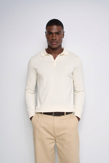 A man wearing a cream long-sleeve polo shirt and beige trousers, standing against a light gray studio background.