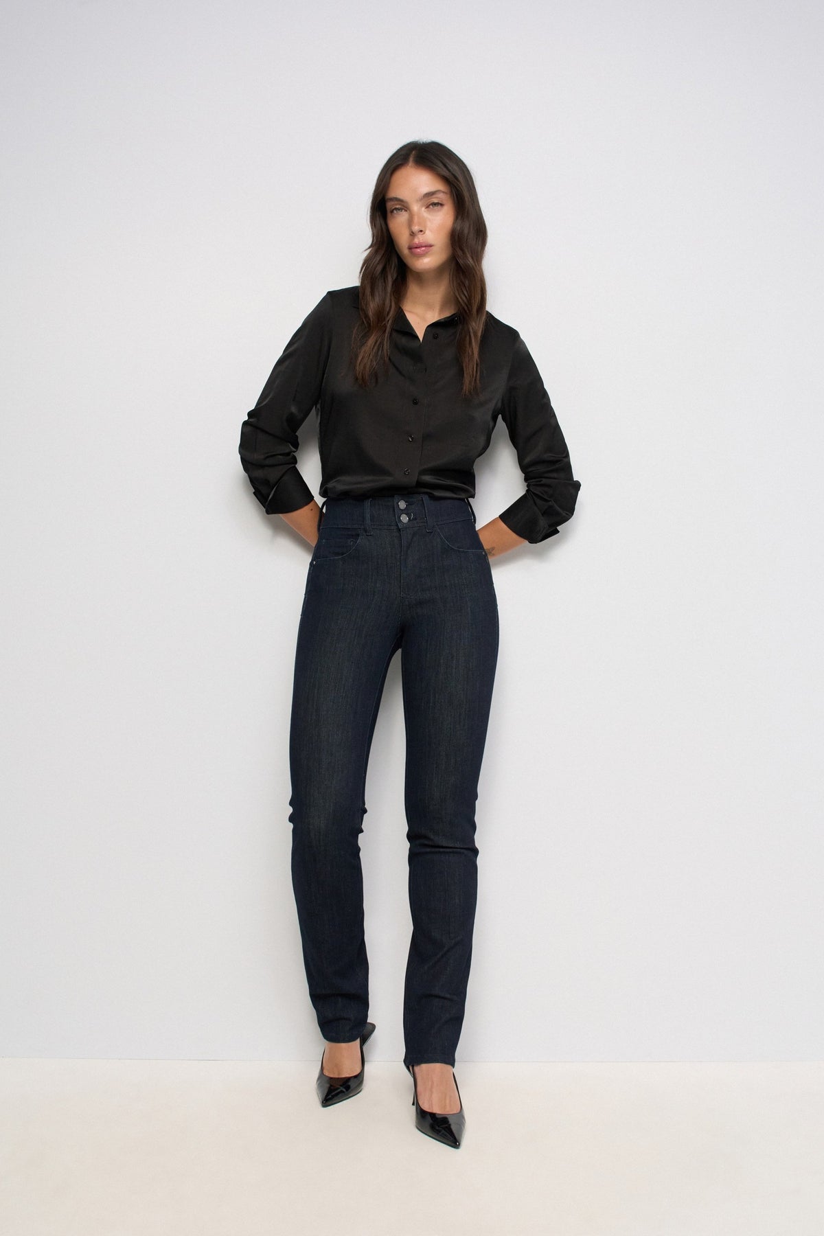 A woman modeling a black long-sleeve satin blouse and dark wash skinny jeans with black pointed-toe heels, standing against a plain white studio background.