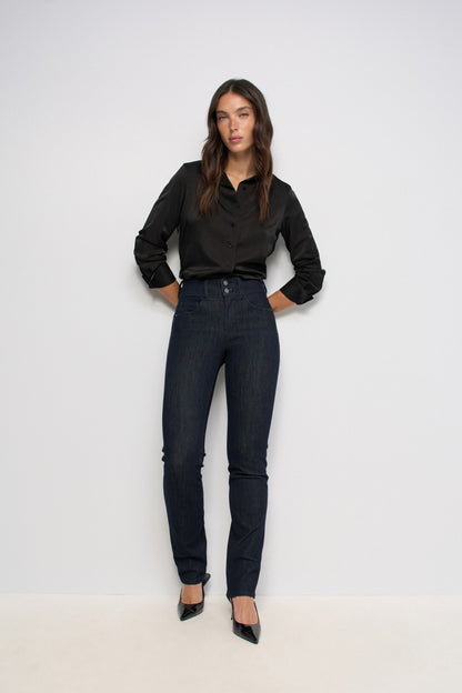 A woman modeling a black long-sleeve satin blouse and dark wash skinny jeans with black pointed-toe heels, standing against a plain white studio background.