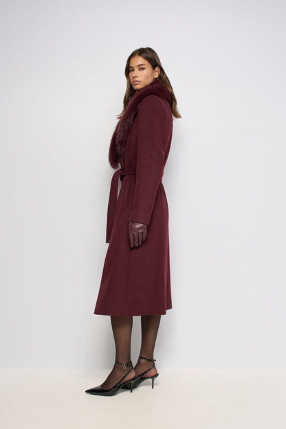 A woman models a long burgundy wool coat with a dark brown fur collar and matching belt, paired with black tights and black slingback heels, against a plain white studio background.