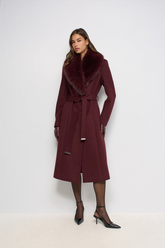 A woman models a long burgundy wool coat with a large dark fur collar, matching belt, and black pointed-toe heels against a plain light gray studio background.