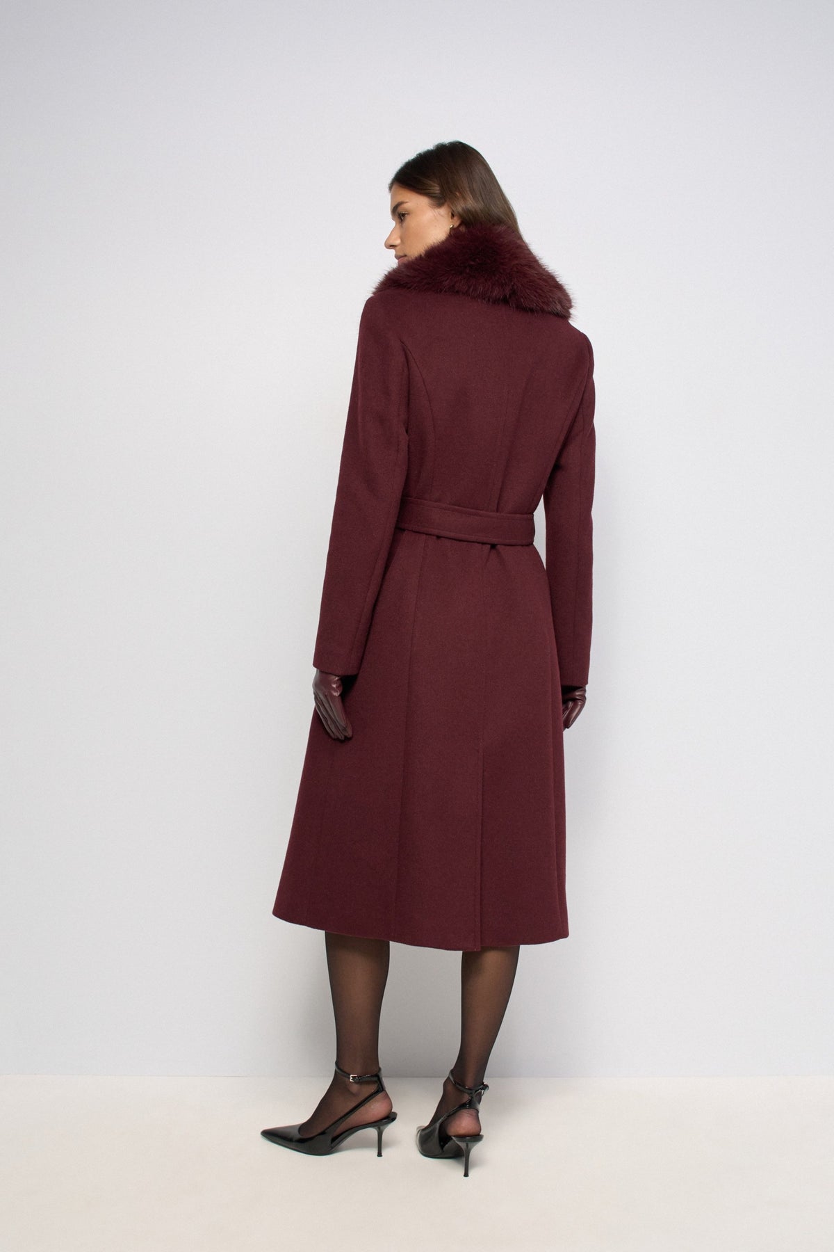 A woman is shown from the back wearing a long burgundy wool coat with a fluffy dark fur collar, paired with black tights and black high heels, against a plain light gray studio background.
