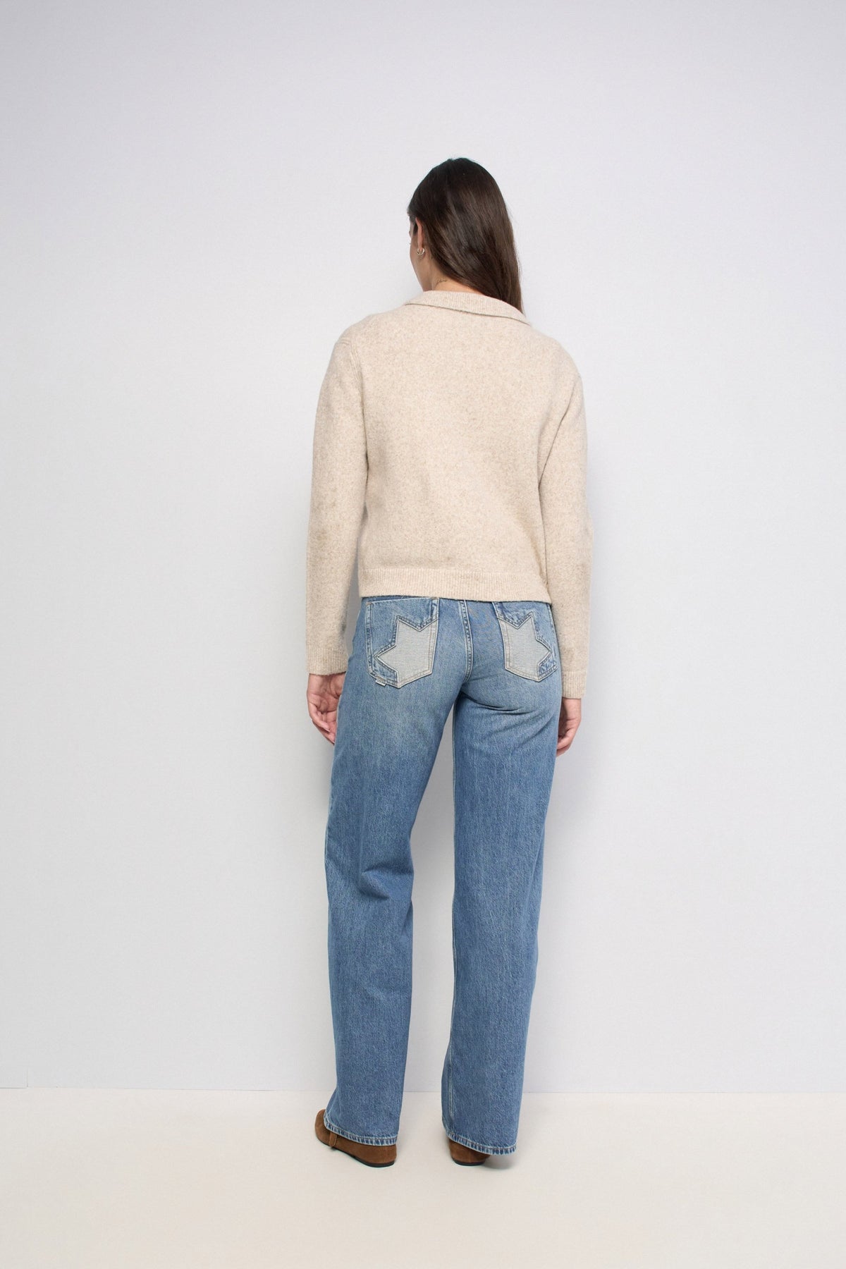 A woman viewed from behind wearing a beige knit sweater and light blue jeans with star-shaped patch details on the back pockets, standing against a plain light gray studio background.
