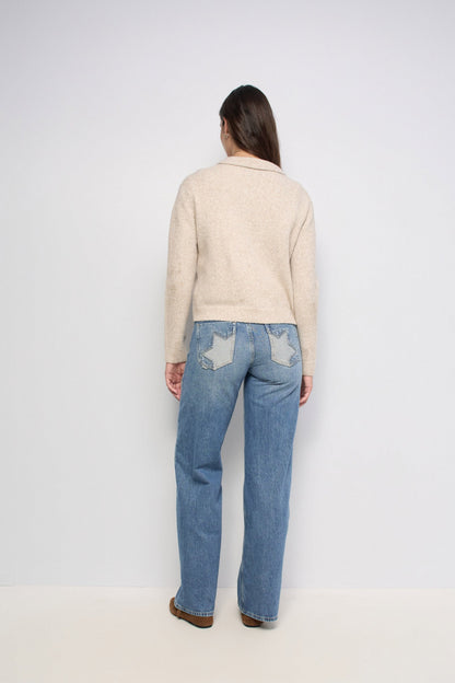 A woman viewed from behind wearing a beige knit sweater and light blue jeans with star-shaped patch details on the back pockets, standing against a plain light gray studio background.