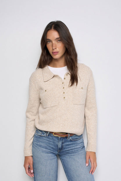 A woman modeling a beige knit polo-style sweater with a buttoned henley collar, chest pockets, and gold-tone buttons, paired with blue jeans and a white undershirt against a light gray studio background.