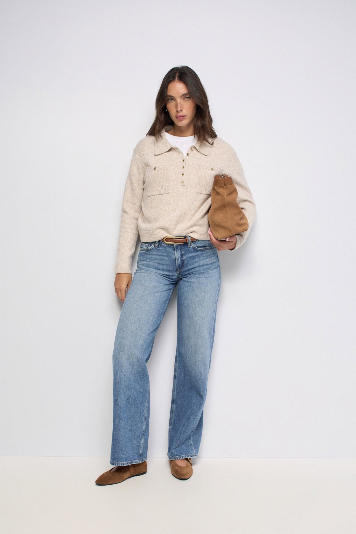 A model stands against a plain light gray studio background wearing a cream knit polo-style sweater over a white top, blue straight-leg jeans, and brown shoes, while holding a brown shoulder bag.