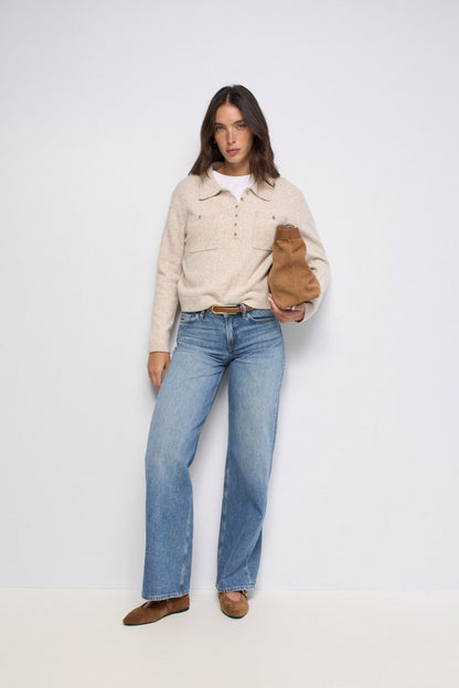 A model stands against a plain light gray studio background wearing a cream knit polo-style sweater over a white top, blue straight-leg jeans, and brown shoes, while holding a brown shoulder bag.