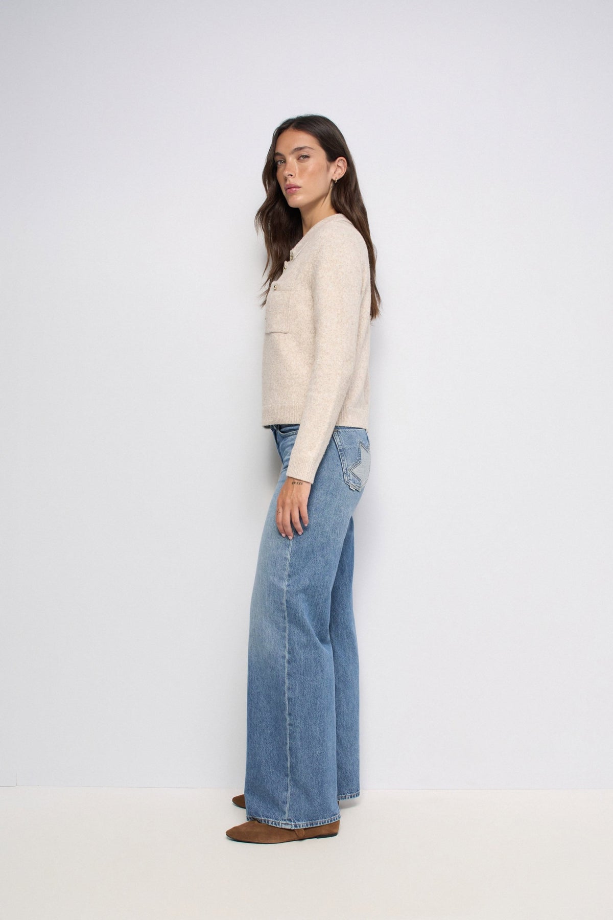 A woman stands in profile wearing a cream-colored knit sweater, wide-leg blue jeans, and brown shoes against a plain white studio background.