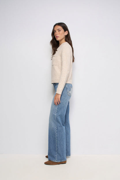 A woman stands in profile wearing a cream-colored knit sweater, wide-leg blue jeans, and brown shoes against a plain white studio background.