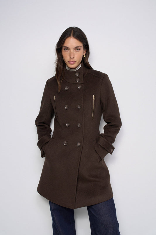 A woman modeling a dark brown double-breasted coat with a high stand collar, button front, and zippered pockets, paired with blue jeans against a light gray studio background.
