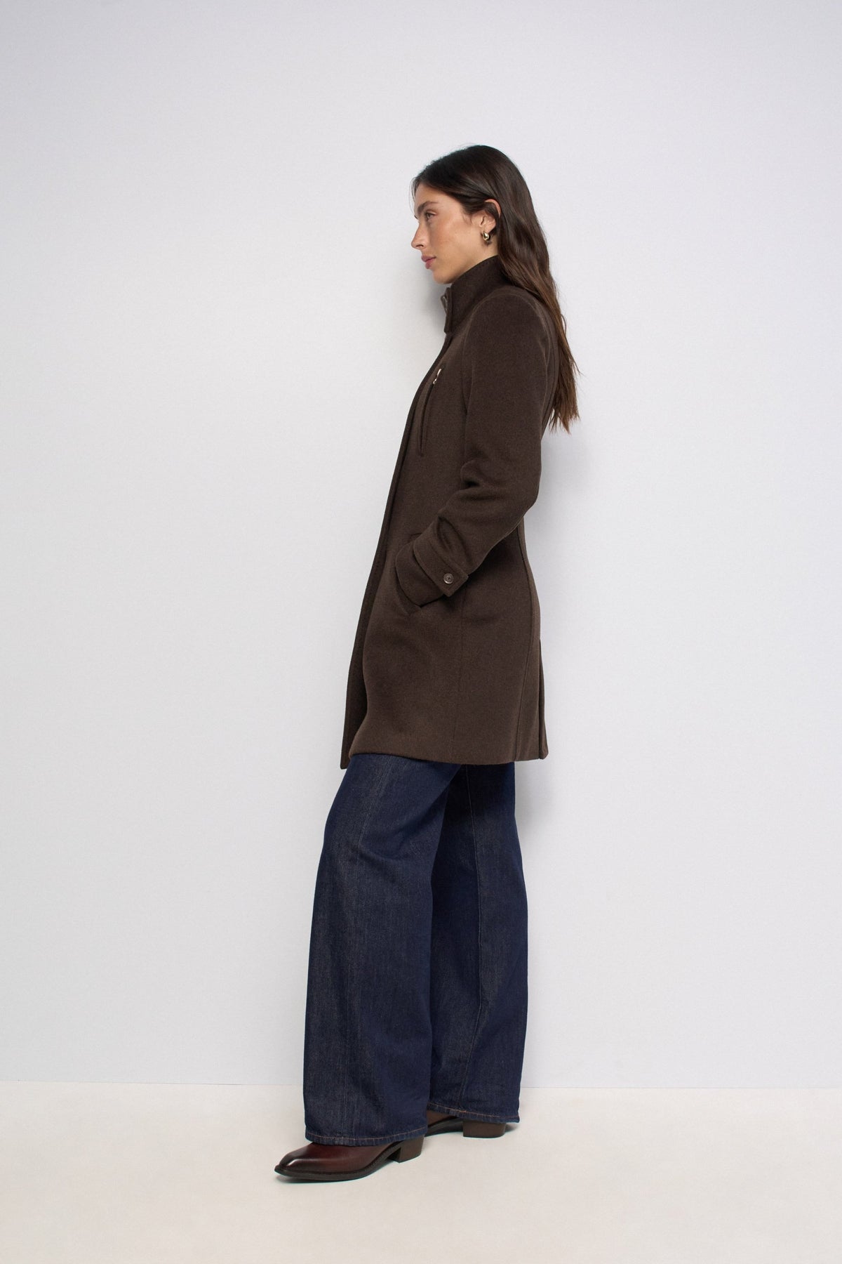 A woman stands in profile wearing a dark brown mid-length coat, dark wide-leg jeans, and brown shoes against a plain light gray studio background.
