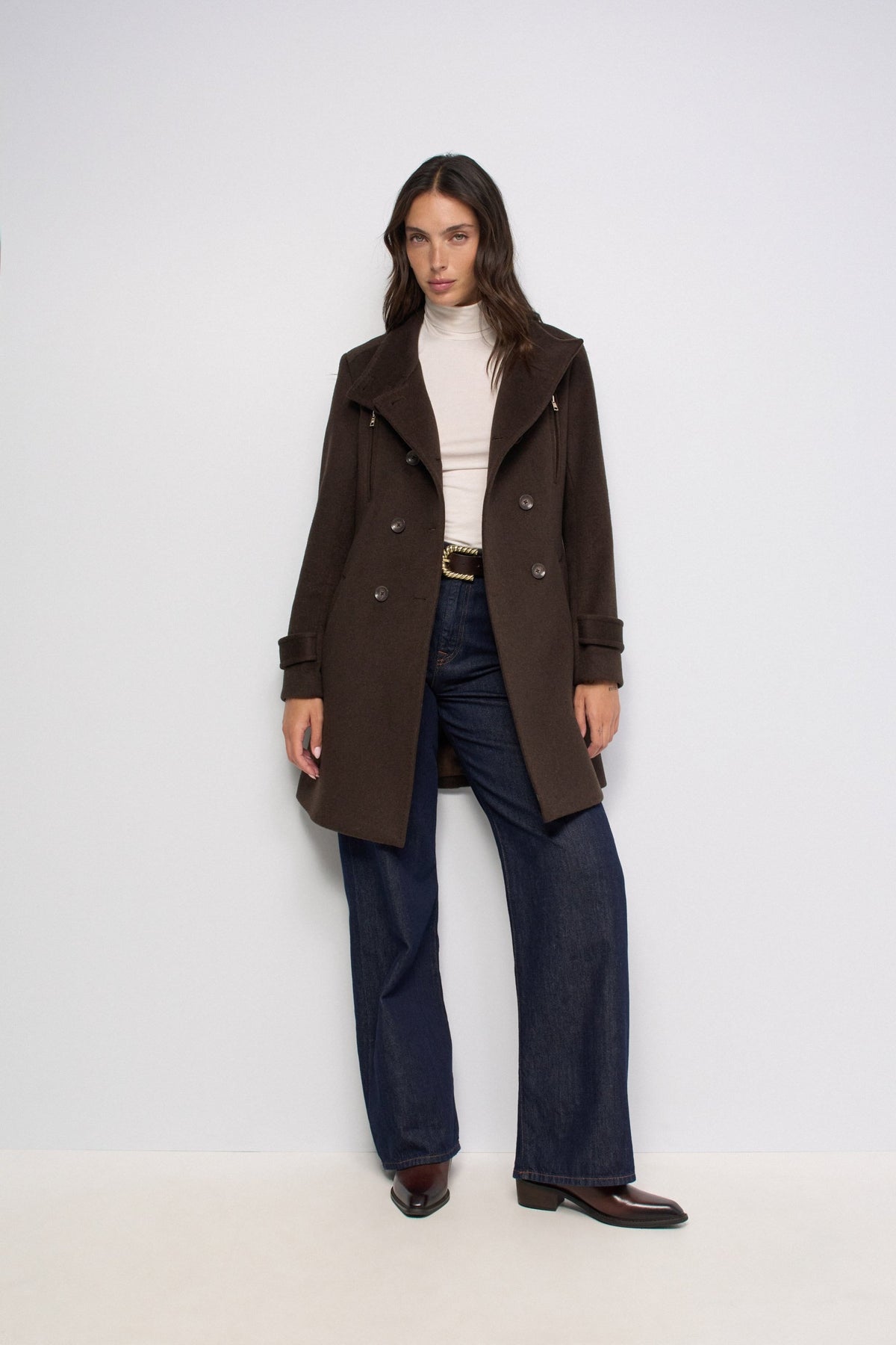 A woman modeling a dark brown double-breasted coat over a cream turtleneck, dark blue wide-leg jeans, and brown leather shoes, standing against a plain white studio background.