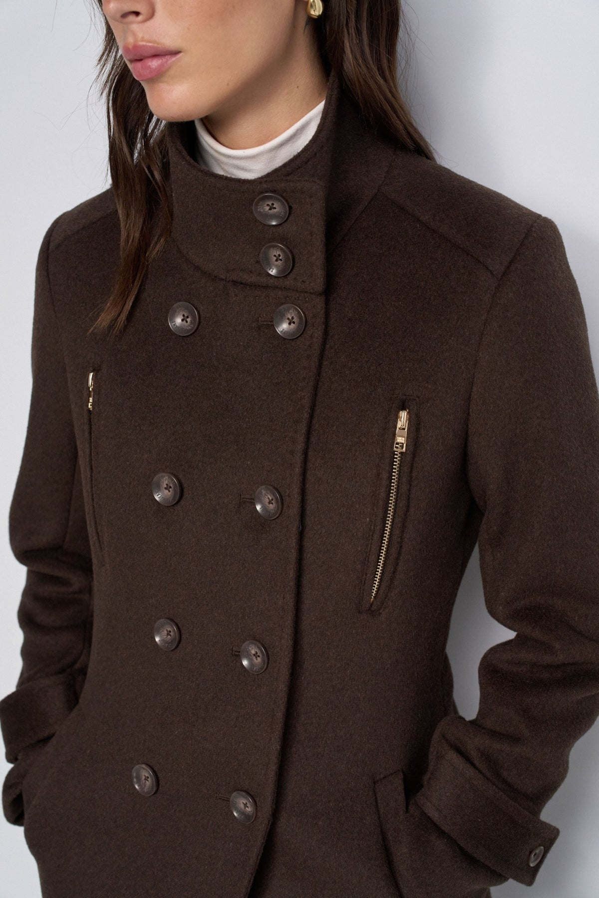 A close-up of a model wearing a dark brown wool-blend coat with a high stand collar, double-breasted buttons, and a vertical zip pocket on the chest.