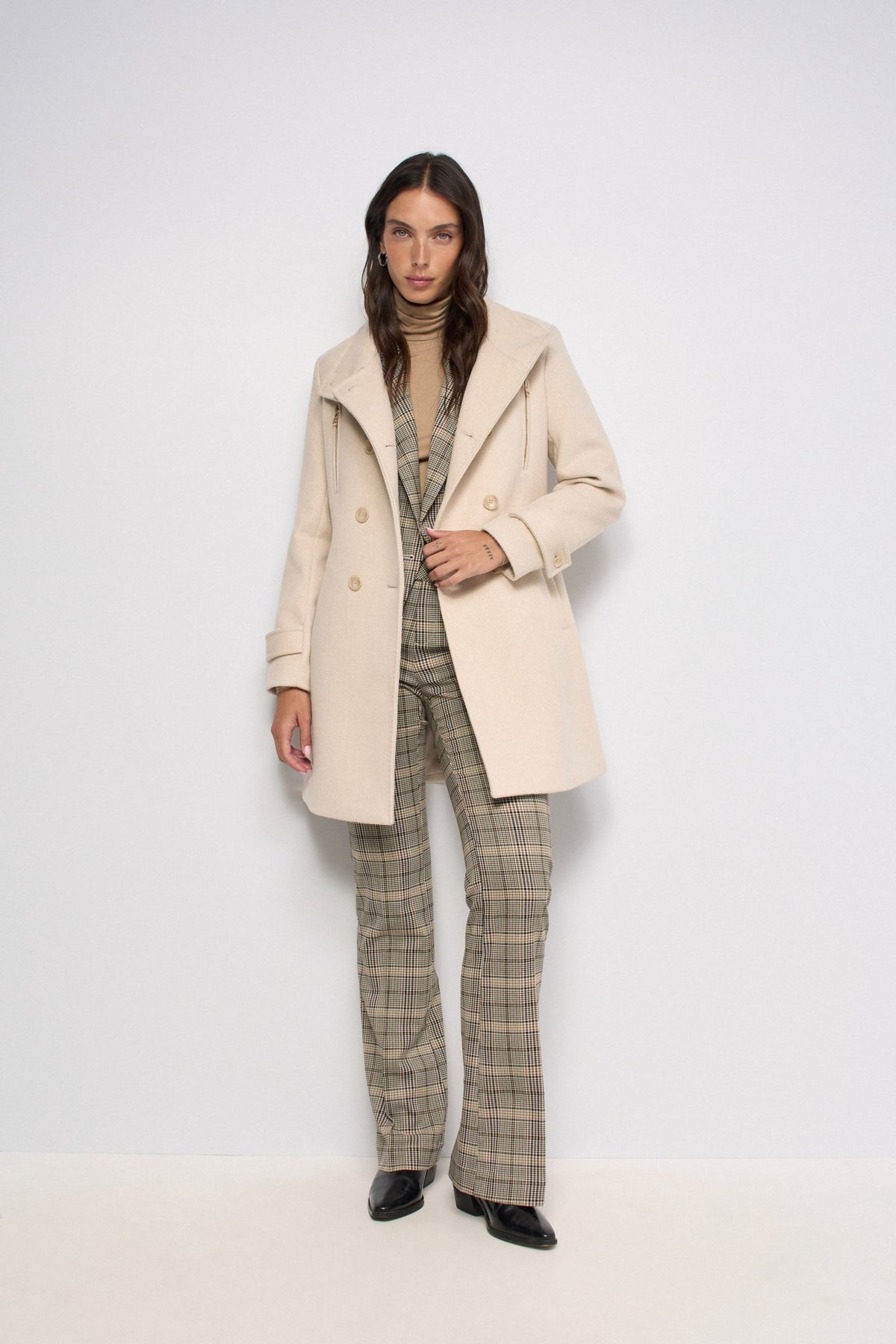 A model stands against a plain white studio background wearing a light beige double-breasted coat over a beige turtleneck, paired with beige and brown plaid trousers and black shoes.