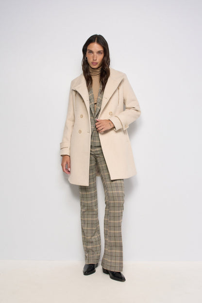 A model stands against a plain white studio background wearing a light beige double-breasted coat over a beige turtleneck, paired with beige and brown plaid trousers and black shoes.