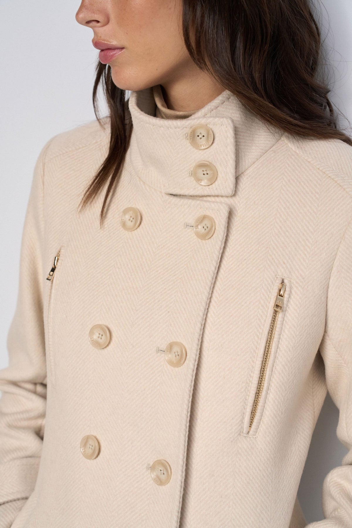 A close-up of a model wearing a cream-colored textured wool coat with a high collar, double-breasted buttons, and a zippered pocket.