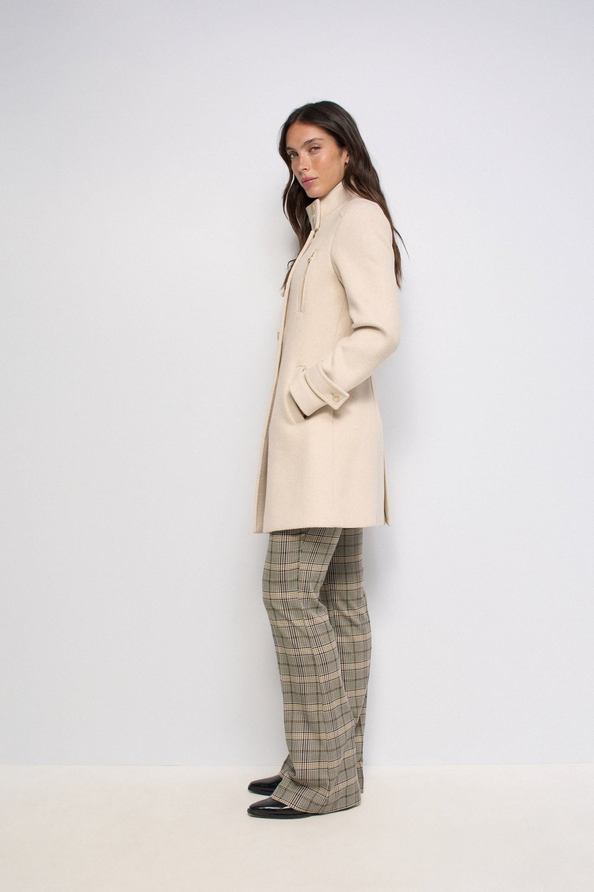 A woman stands in profile wearing a long cream-colored coat, beige plaid trousers, and black shoes against a plain light gray studio background.