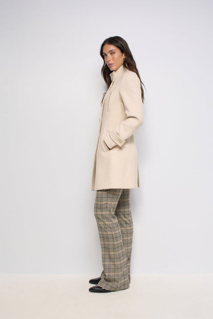 A woman stands in profile wearing a long cream-colored coat, beige plaid trousers, and black shoes against a plain light gray studio background.