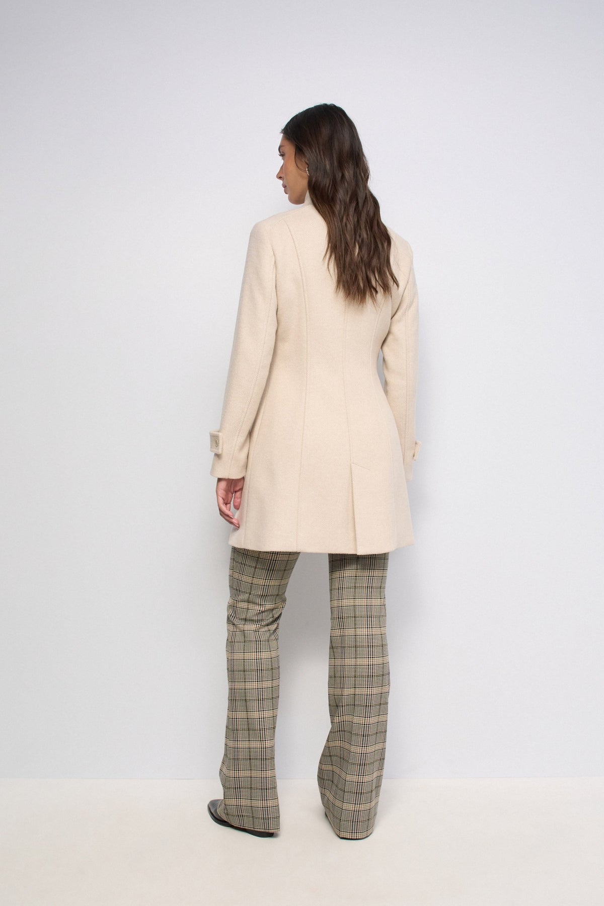 A woman is shown from the back wearing a light beige mid-length blazer and olive green plaid trousers, standing against a plain white studio background.
