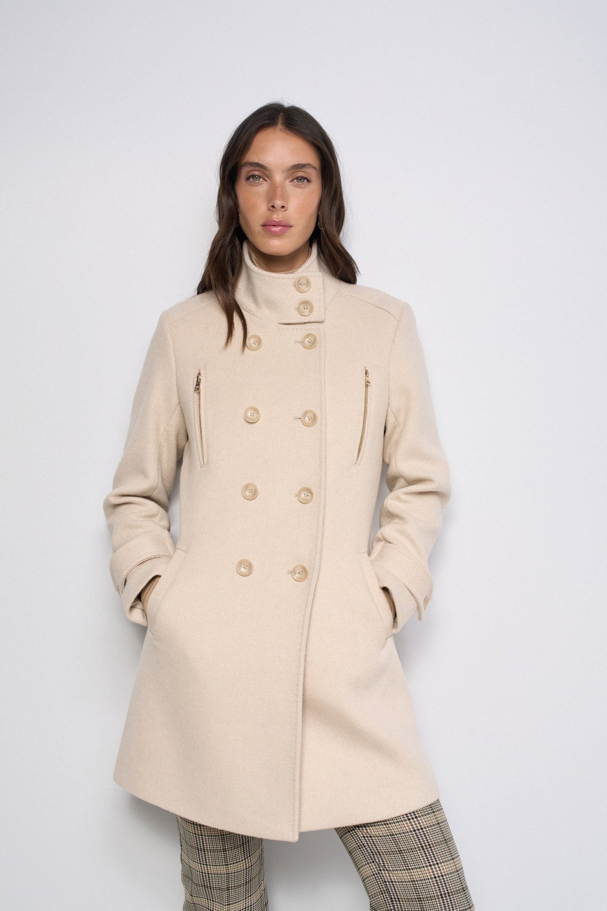 A woman modeling a mid-length cream-colored double-breasted coat with gold-tone buttons and zippered pockets, paired with plaid trousers, against a plain light gray studio background.