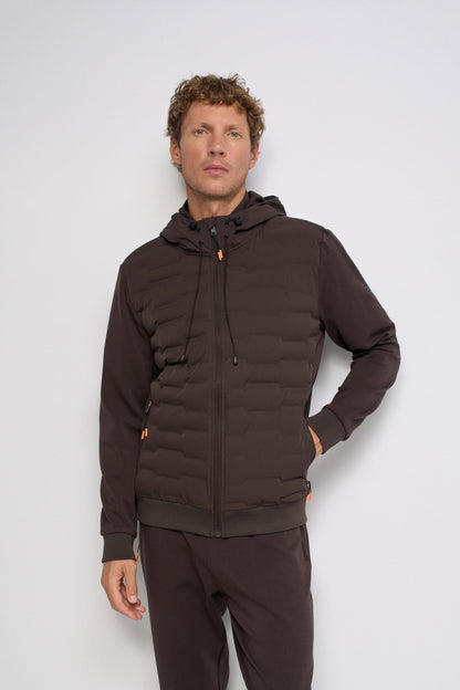 A man wearing a dark brown hooded puffer jacket with a full zip and drawstrings, paired with matching dark brown sweatpants, standing against a light gray studio background.