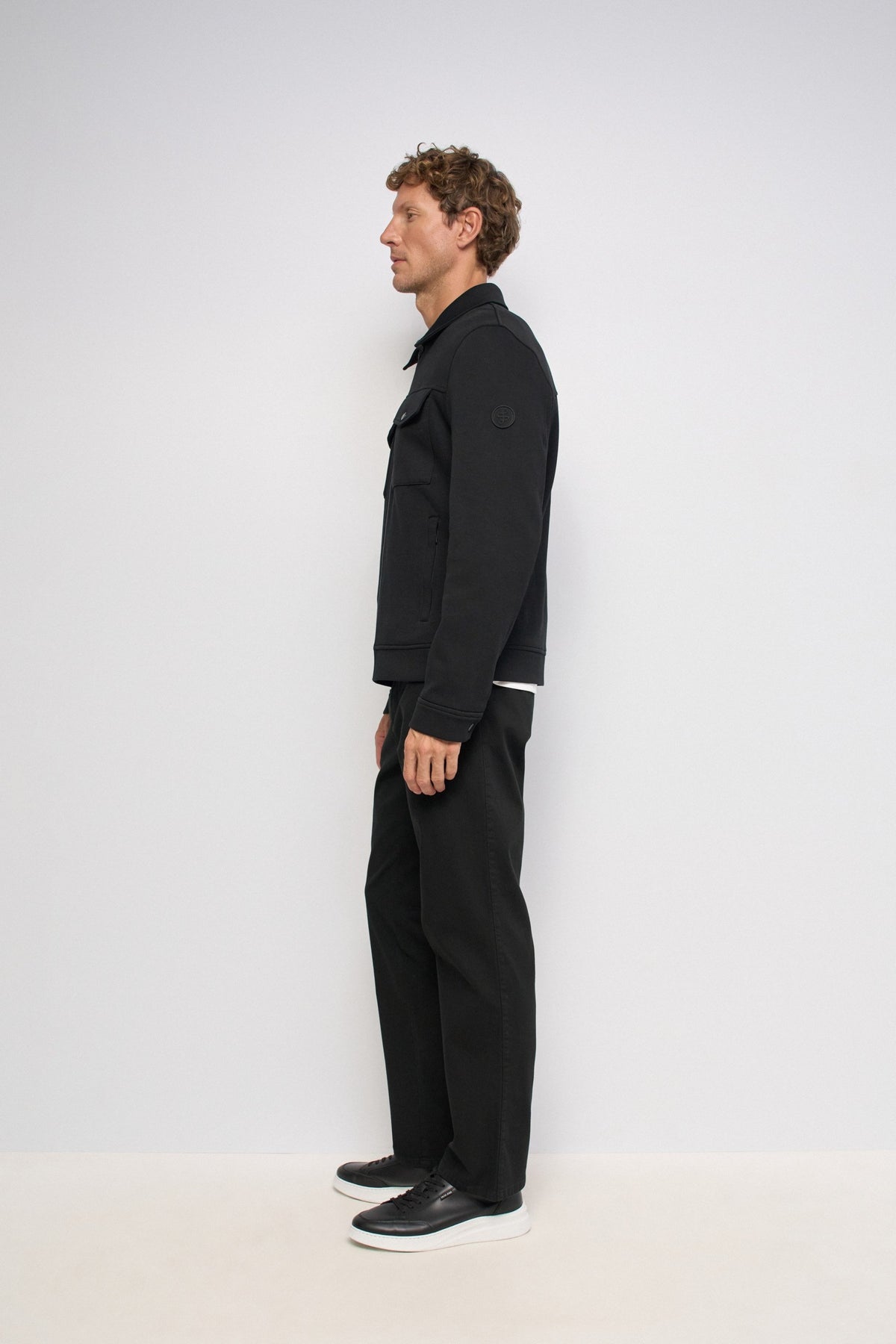 A man wearing a black zip-up jacket and black pants, standing against a light gray studio background.