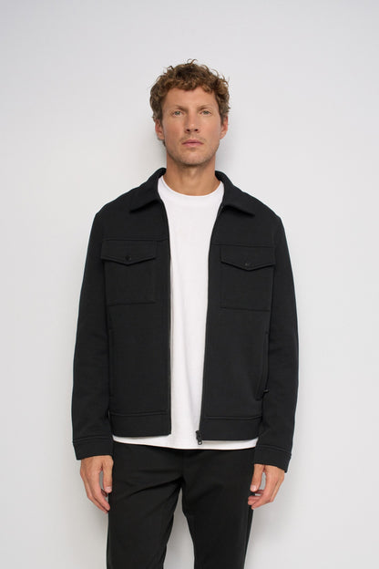A man wearing a black zip-up jacket over a white t-shirt and black pants, standing against a light gray studio background.