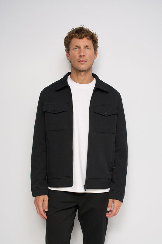 A man wearing a black zip-up jacket over a white t-shirt and black pants, standing against a light gray studio background.