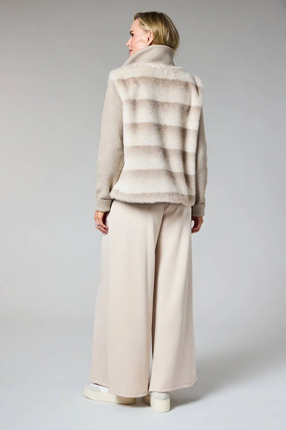 A model shown from the back wearing a beige and taupe striped faux fur vest over a beige knit sweater and wide-leg beige trousers, set against a plain light gray studio background.