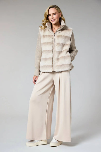 A woman modeling a beige faux-fur zip-front vest over a beige long-sleeve top and wide-leg beige trousers, standing against a light gray studio background.