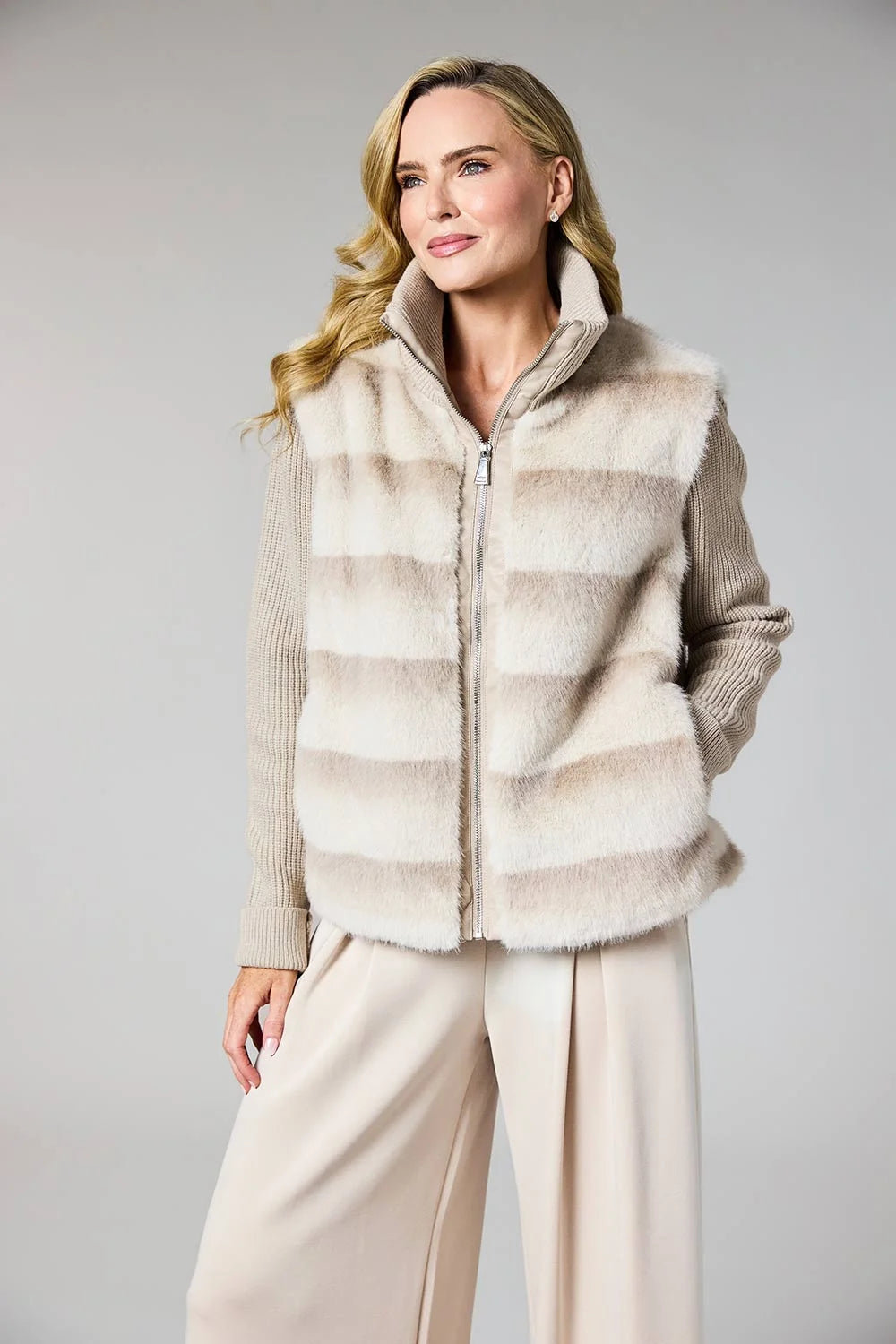 A woman modeling a beige and cream faux fur vest over a ribbed knit sweater and cream wide-leg trousers, photographed against a neutral light gray studio background.