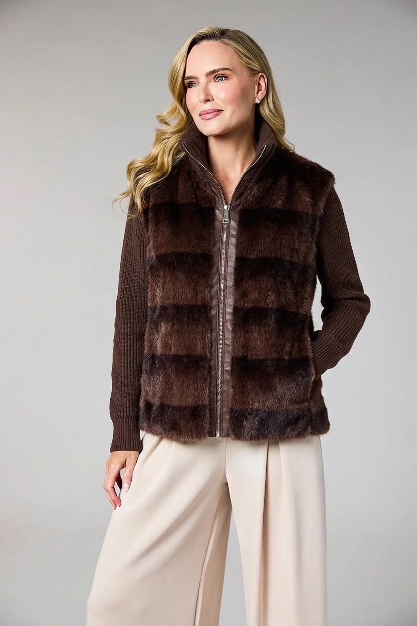 A woman modeling a brown faux fur vest with horizontal stripes over a dark brown ribbed sweater and cream wide-leg pants, against a solid gray studio background.
